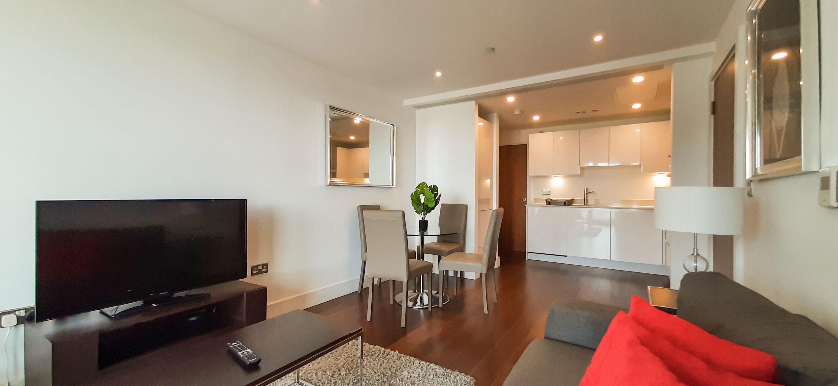 A modern living room with a flat-screen TV, a coffee table, and a sofa with red cushions. There is a dining area with a table and chairs, and a kitchen in the background.