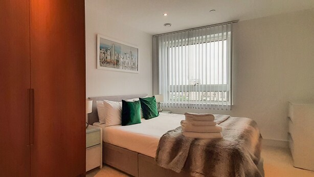 A modern bedroom with a bed, a nightstand, a window with blinds, and a cityscape painting on the wall.