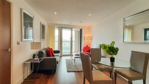 A modern living room with a sofa, a glass table, and a chair. There is a large window and a door leading to another room.
