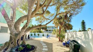 Un patio trasero con un árbol grande, una piscina y una cerca blanca.