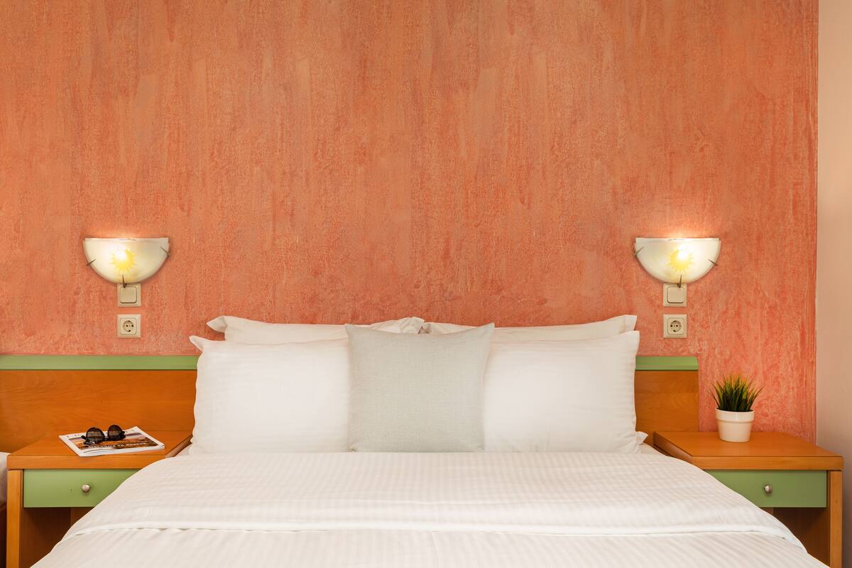 A neatly made bed with white linens, a green nightstand with a magazine and sunglasses, and a small potted plant on the nightstand.