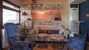 A cozy living room with a stone wall, a cityscape painting, a sofa, and blue upholstered chairs. - Dias Hotel & Spa (Dio-Olympos)