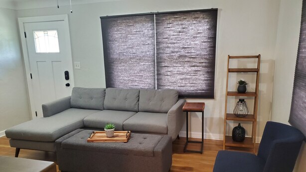 A living room with a grey sectional sofa, a small wooden table with a tray and plant, a blue armchair, and a wooden shelf with decorative items.