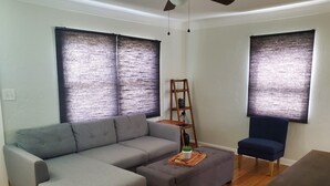 A living room with a grey sectional sofa, a small ottoman, a wooden ladder shelf with plants, and a dark blue armchair.