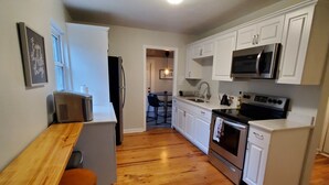 A compact kitchen with white cabinets, a stainless steel refrigerator, a microwave, and a stove. There is a wooden dining table with two chairs.