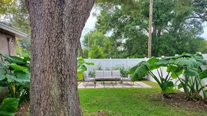 A backyard with a large tree, a white fence, and a patio with outdoor furniture.