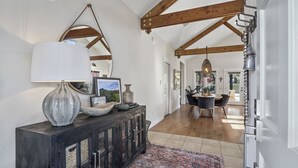 Un pasillo amplio con una consola de madera oscura, un espejo grande y una lámpara colgante. La habitación cuenta con vigas de madera a la vista y una vista hacia un comedor con una mesa y sillas.