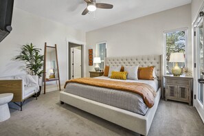 Een slaapkamer met een groot bed, een plafondventilator, een nachtkastje met een lamp, een potplant en uitzicht op de omgeving door een raam.