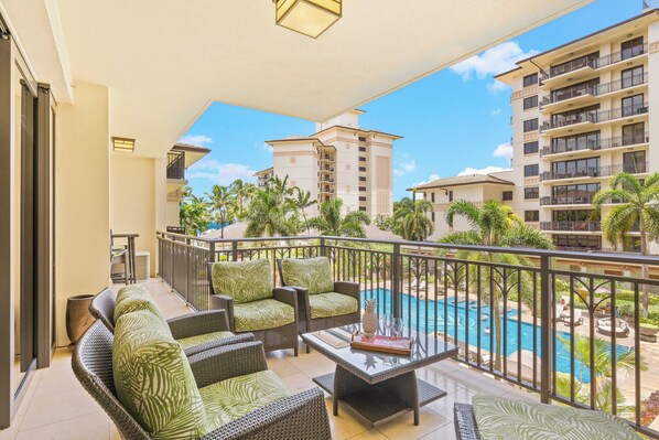 A balcony with wicker furniture, a glass table, and a view of a swimming pool and surrounding buildings.