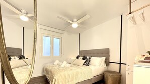 Une chambre à coucher avec un lit, un ventilateur de plafond, une fenêtre, un miroir, une poubelle et un petit tapis.