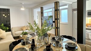 Un espace repas moderne avec une table ronde en bois, dressée pour six personnes, entourée de chaises. On aperçoit un canapé et une table basse agrémentée d’objets décoratifs à l’arrière-plan.