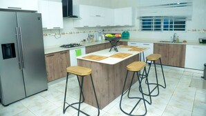 A modern kitchen with a central island, bar stools, a refrigerator, and a window with blinds. - Spacious 5 BR Villa with amazing views Bar&Pool | family & large groups | Wi-Fi (Accra)