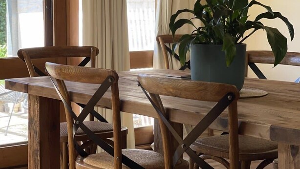 Mesa de jantar de madeira com cadeiras, um vaso de planta e vista para o exterior por grandes janelas.