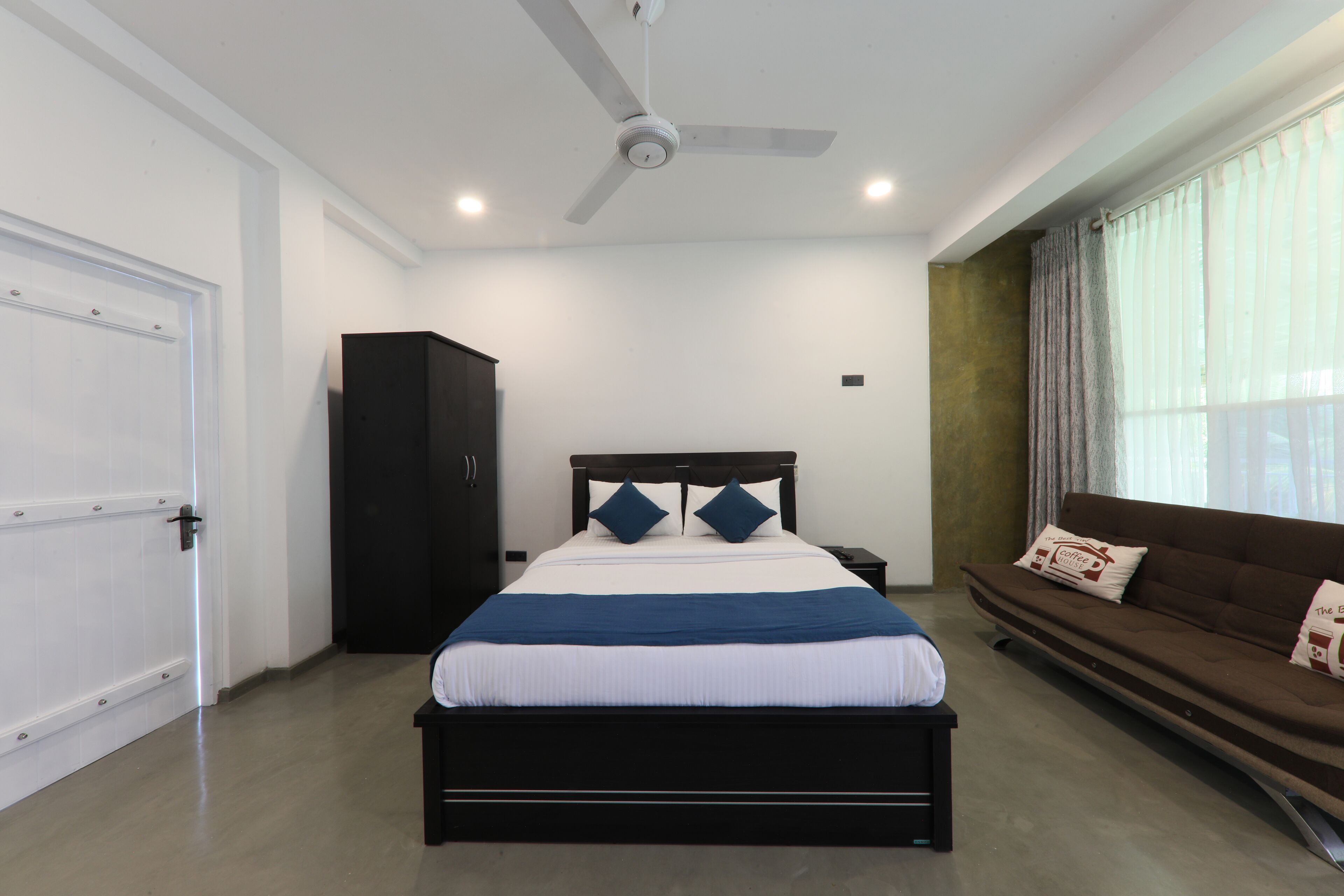 A bedroom with a bed, a ceiling fan, a wardrobe, a sofa, and a window with curtains.