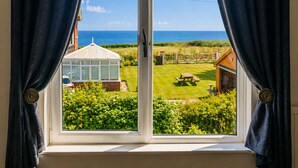 A window with curtains, a view of a garden, a conservatory, and a house with a picnic table. - Spacious 12-Bedroom Coastal Retreat Private Annex (Mundesley)