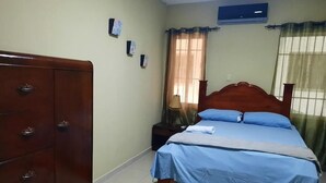 Quarto com cômoda de madeira, cama com roupa de cama azul, mesa de cabeceira, luminária e janela com cortinas.