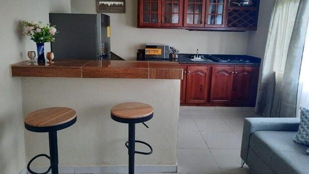 Uma cozinha compacta com uma ilha central, dois bancos, armários de madeira e um sofá cinzento.