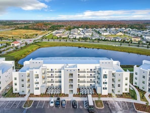A modern multi-story building with a parking lot in front, a large body of water behind it, and a residential area in the background.
