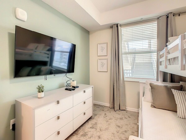 A bedroom with a TV mounted on the wall, a dresser with drawers, a bed with a headboard, and a window with curtains.