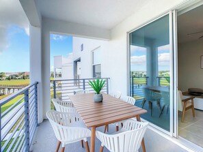 A balcony with a wooden table and white chairs, a potted plant, and a view of a modern building and landscape.