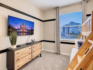 A hotel room with a large flat-screen TV mounted on the wall, a wooden dresser, a bed with a wooden headboard, and a window with blinds.