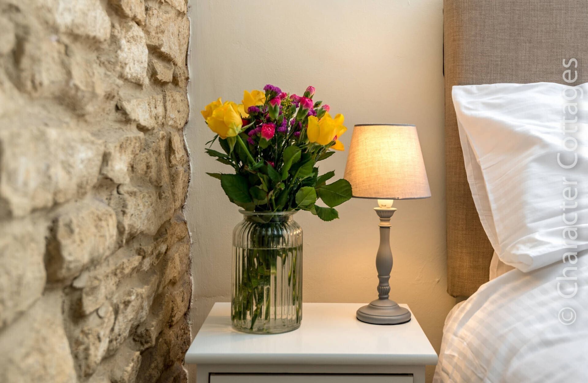 Ein Nachttisch mit einer Glasvase, die Blumen enthält, einer Lampe und einem Bett mit einem weißen Kissen.
