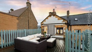 A patio with a wicker sofa and a table, a wooden deck, and a white picket fence. - Eastnor (Moreton-in-Marsh)