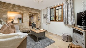 A cozy living room with a stone wall, a wooden coffee table, a sofa, a television, and a window with curtains. - Eastnor (Moreton-in-Marsh)