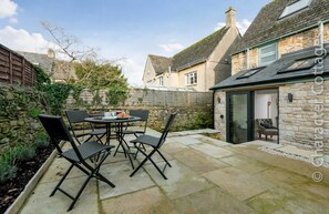 Une terrasse avec une table ronde et quatre chaises, un mur en pierre et une maison avec une porte en verre et une chaise à l’intérieur.