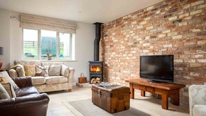A living room with a brick wall, a fireplace, a sofa, a leather chair, a television, and a wooden table. - Hetty's Stables (Cheltenham)
