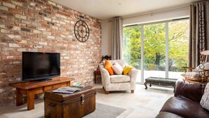 A living room with a brick wall, a television, a sofa, a chair, and a large window with a view of trees. - Hetty's Stables (Cheltenham)