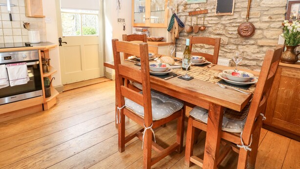 A cozy dining area with a wooden table set for four, surrounded by wooden chairs. A chandelier hangs above, and the walls are adorned with various decorative items and a stone wall. - Carter's Cottage (Cheltenham)