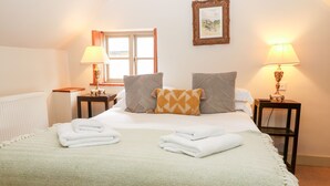 A neatly made bed with folded towels, two bedside lamps, a framed picture on the wall, and a window with blinds. - Carter's Cottage (Cheltenham)
