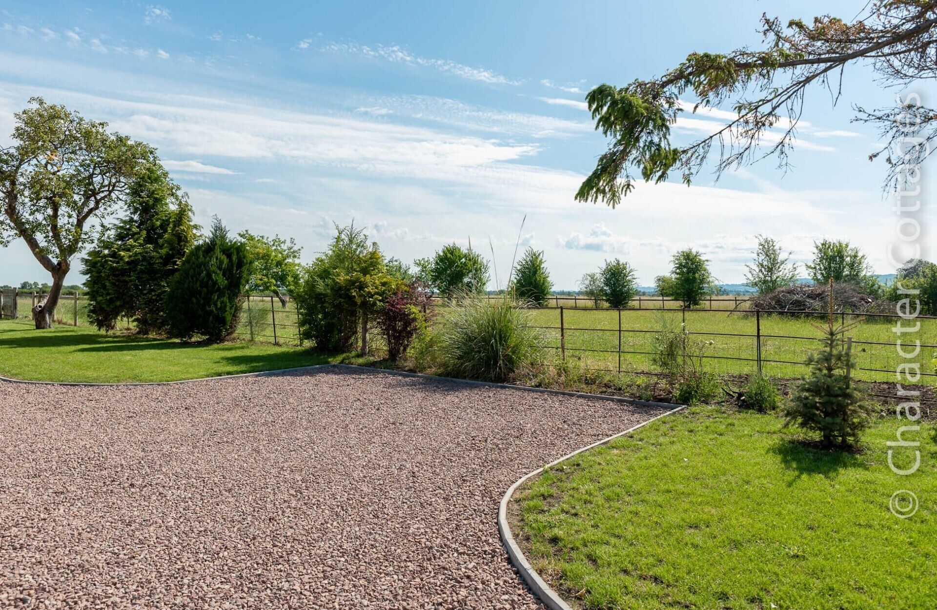 A gravel driveway leading to a grassy area with a fence and trees in the background.