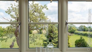Una ventana con tres vasos de agua y una botella sobre una mesa, con vistas a un paisaje verde con árboles y una valla.