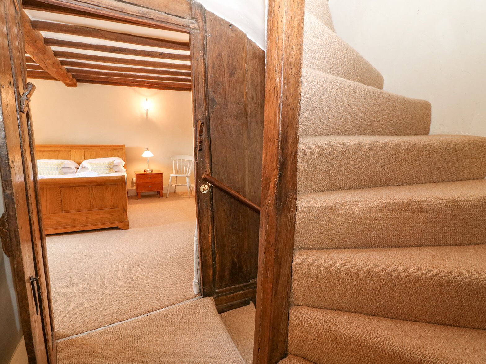 A bedroom with a wooden bed, a nightstand, a chair, and a staircase leading to another room.