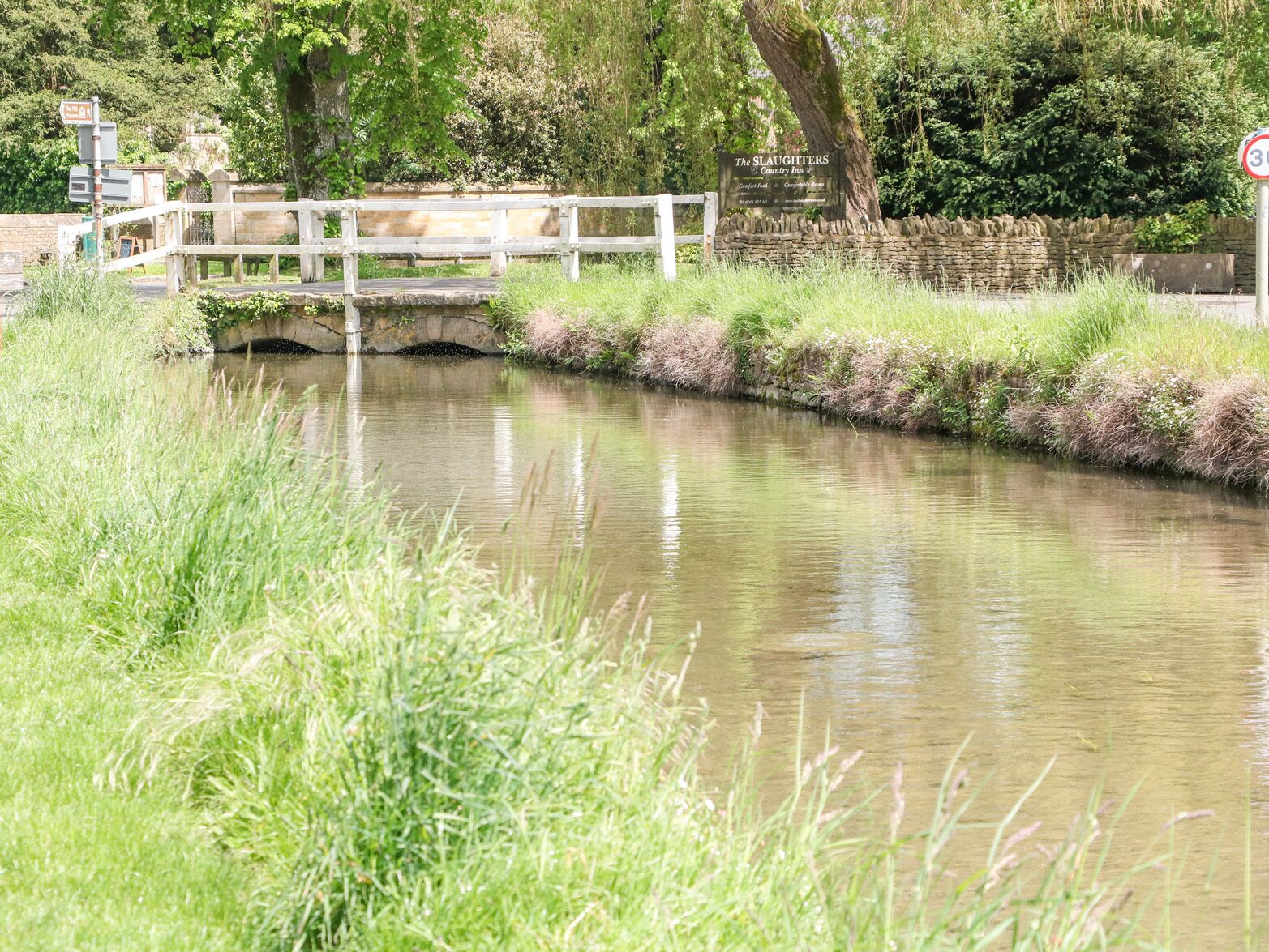 Ein Kanal mit einer Holzbrücke und einem Schild für The Slaughters Country Inn.