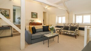 A spacious loft with a high ceiling, a grey sofa, a floral armchair, and a dining table. - The Apartment (Cheltenham)