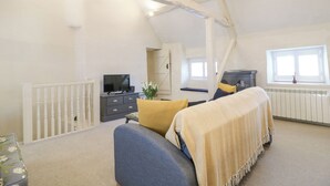 A spacious attic bedroom with a high ceiling, a bed with a beige blanket, a television on a dresser, and a window with a view. - The Apartment (Cheltenham)