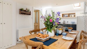 A dining area with a wooden table set for four, a vase of flowers, and a kitchen in the background. - Copperbeech (Cheltenham)