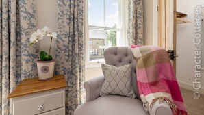 A bedroom with a window, a chair with a blanket, a bedside table with a potted plant, and floral curtains. - Copperbeech (Cheltenham)