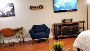Een moderne woonkamer met een blauwe fauteuil, een houten salontafel met een plant, een televisie aan de muur en een klein rekje met decoratieve objecten.