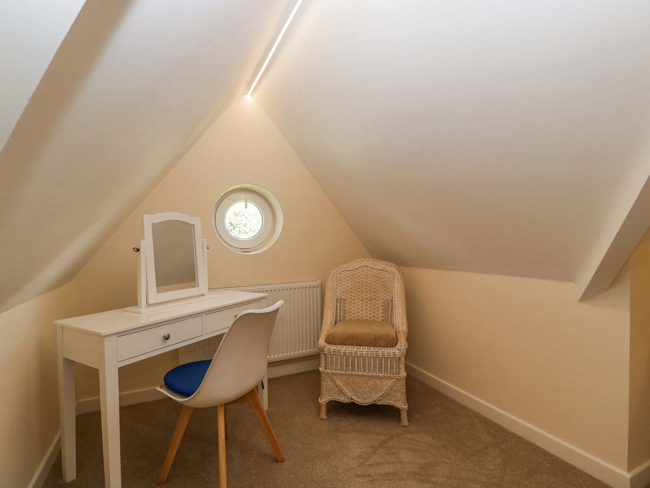 A small, triangular attic bedroom with a white desk, a chair, a wicker chair, a round window, and a radiator.