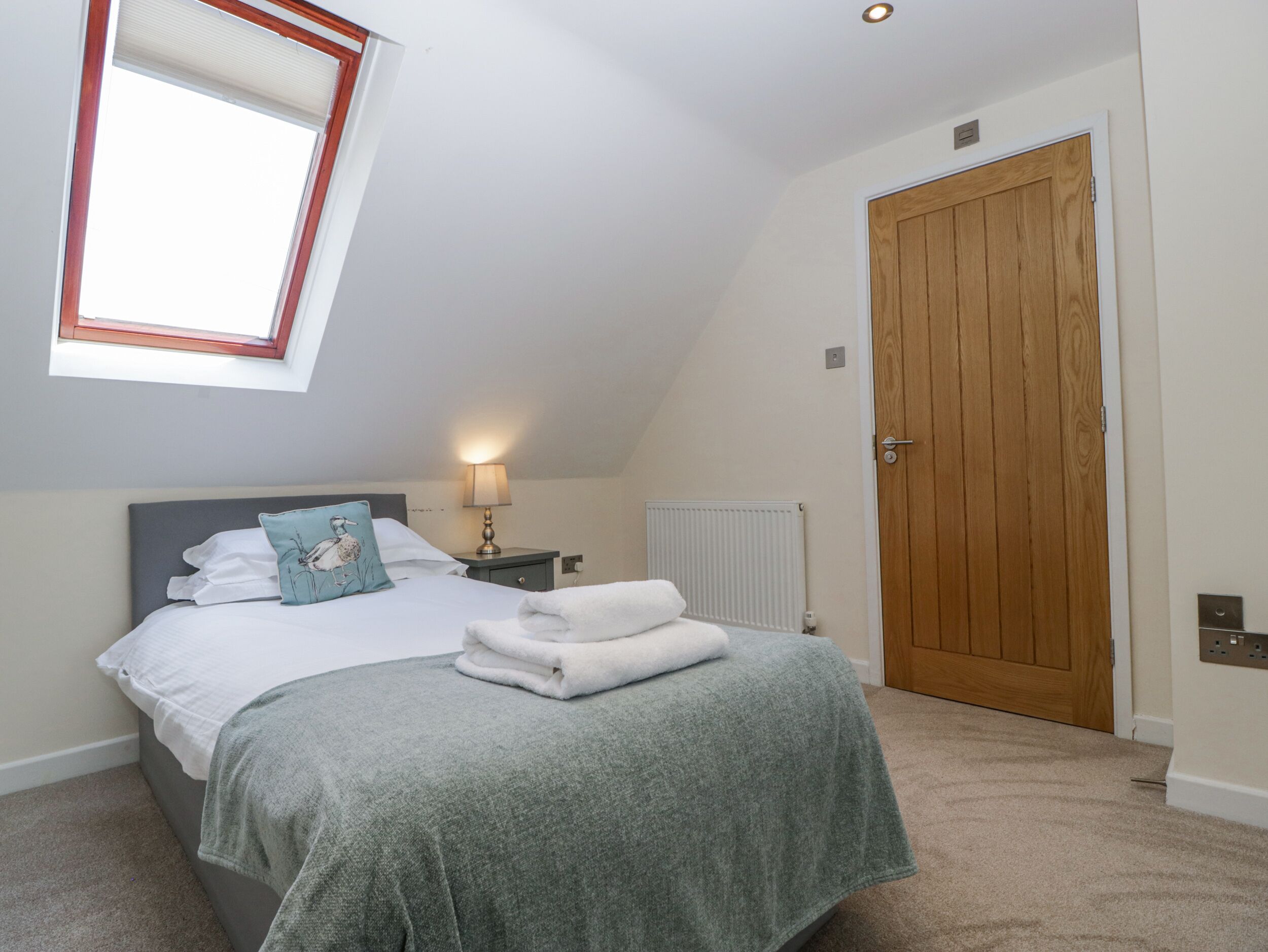 A bedroom with a bed, a nightstand, a lamp, a radiator, a wooden door, and a skylight.