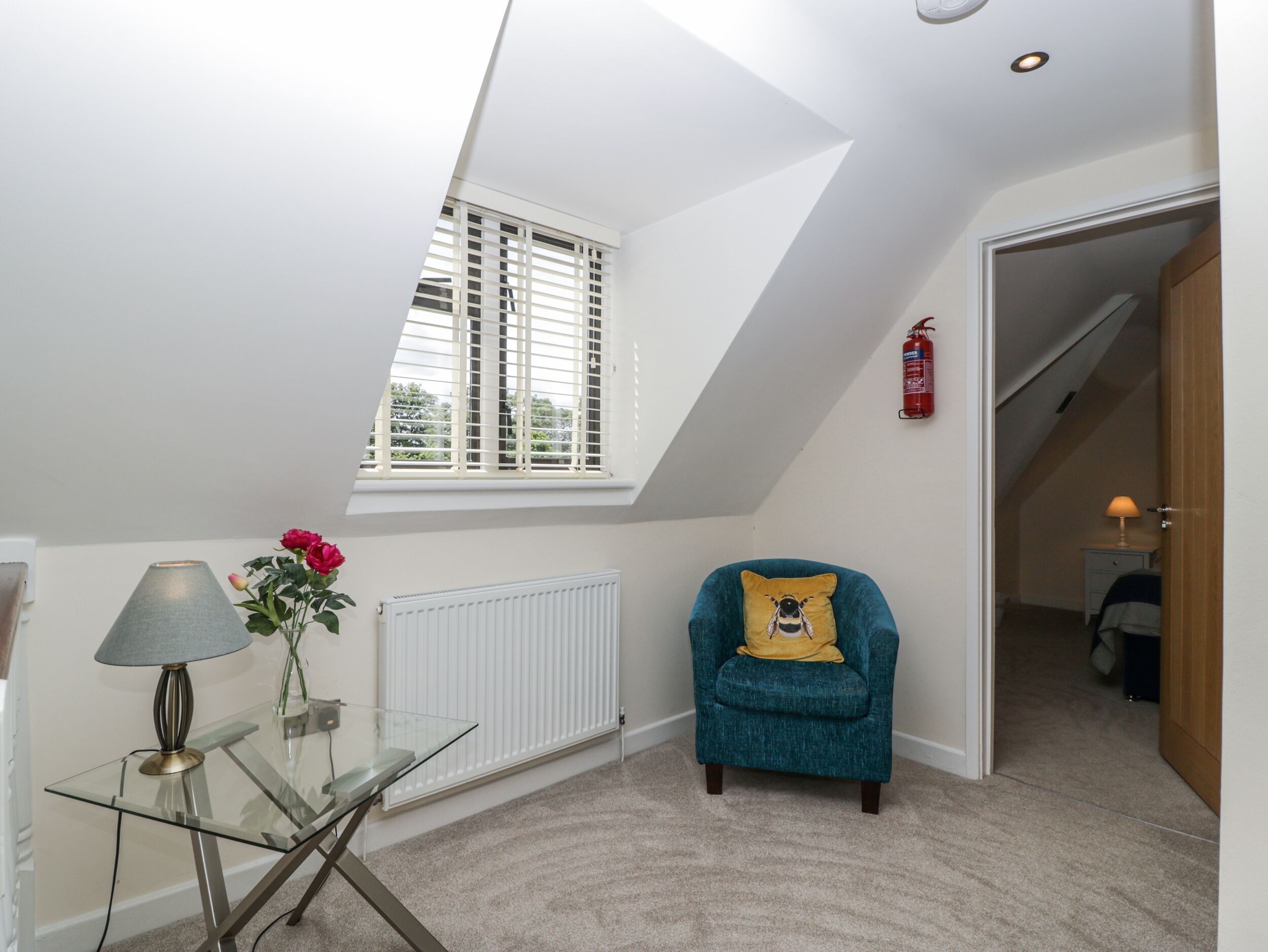 A room with a sloped ceiling, a window with blinds, a chair with a pillow, a glass table with a lamp, and a radiator.