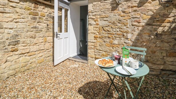 A stone building with a door open to a gravel area. A table with plates, a carton of Tropicana juice, and a mug is set outside. - The Bath House (Moreton-in-Marsh)