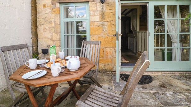 A wooden table set with breakfast items, a stone wall, and a green door leading to a stable. - Red Fawn Cottage (Moreton-in-Marsh)