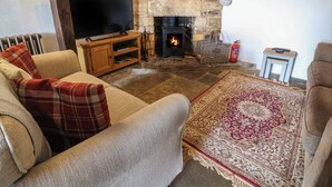 A cozy living room with a stone fireplace, a television on a wooden stand, a sofa with plaid cushions, and a patterned rug. - Red Fawn Cottage (Moreton-in-Marsh)