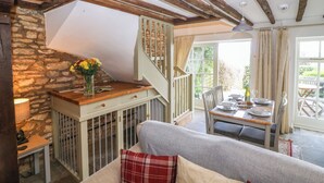A cozy room with a stone wall, wooden beams, a dining table set for two, and a staircase leading to an upper level. - Red Fawn Cottage (Moreton-in-Marsh)