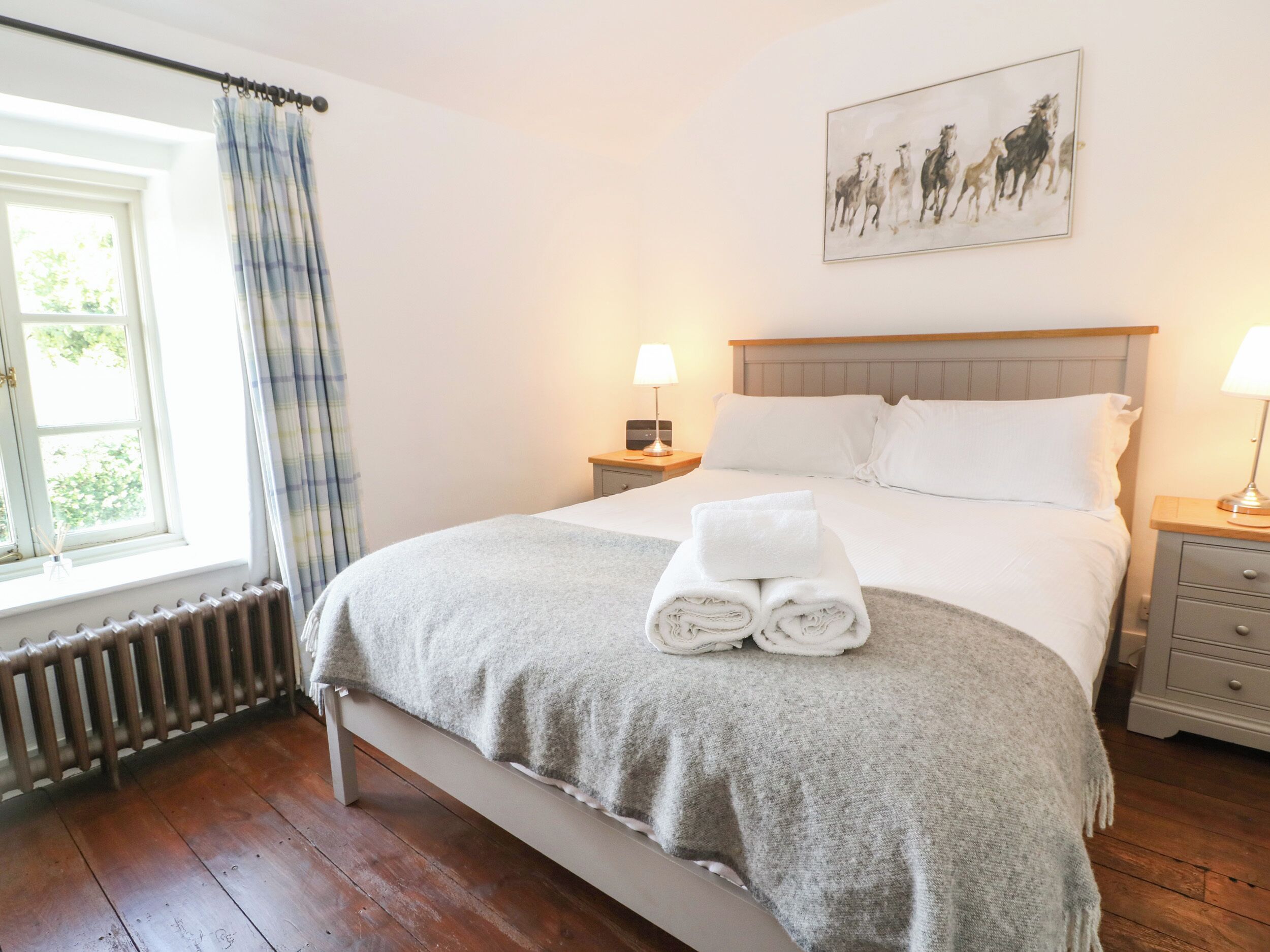Una cama bien hecha con una manta gris y toallas enrolladas, dos mesitas de noche con lámparas, una ventana con cortinas y un cuadro de caballos en la pared.